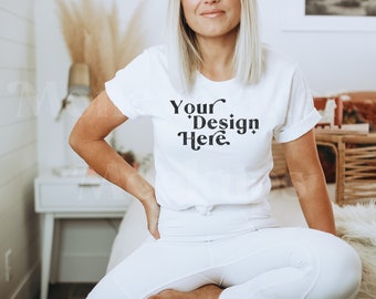 Peut inclure: Un t-shirt blanc avec le texte "Your Design Here" imprimé en noir sur le devant. Le t-shirt est porté par une personne assise sur un lit blanc.