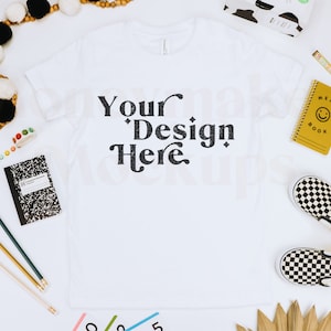 K&ouml;nnte beinhalten: Wei&szlig;es T-Shirt mit dem Text "Your Design Here" in schwarzer Schrift. Das Shirt liegt flach auf einer wei&szlig;en Oberfl&auml;che mit Schulutensilien, einem Notizbuch und einem Paar karierten Slipper.