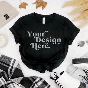 May include: Black t-shirt with the text "Your Design Here" in a white, vintage-style font. The shirt is laid flat on a white surface with a beige beanie, white pumpkin, plaid shirt, sunglasses, white boots, and dried pampas grass.