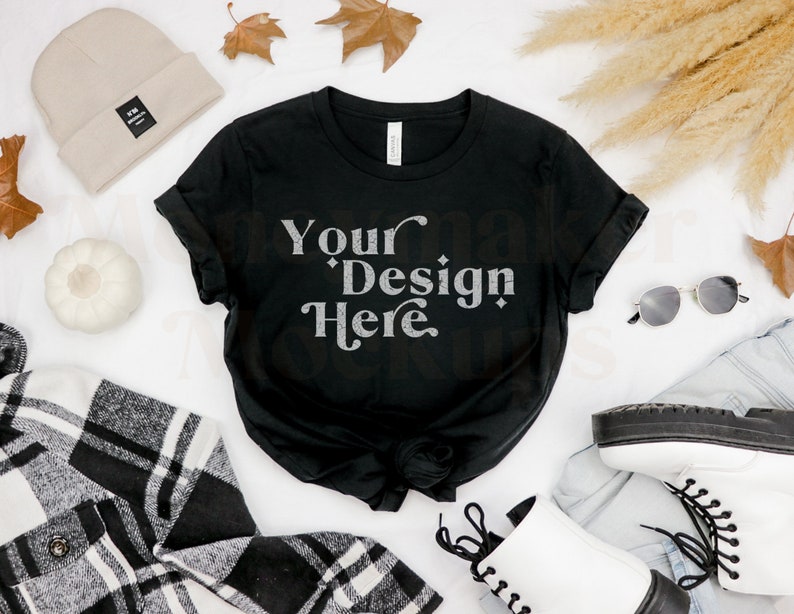 May include: Black t-shirt with the text "Your Design Here" in a white, vintage-style font. The shirt is laid flat on a white surface with a beige beanie, white pumpkin, plaid shirt, sunglasses, white boots, and dried pampas grass.
