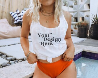 May include: A white tank top with the text "Your Design Here" printed on the front. The tank top is worn by a person with long blonde hair and is tied at the waist. The person is standing near a pool.