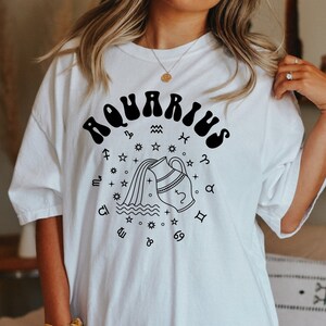 Puede incluir: Camiseta blanca de gran tama&ntilde;o con un gr&aacute;fico negro del signo astrol&oacute;gico de Acuario. El gr&aacute;fico presenta una jarra de agua estilizada que vierte agua y est&aacute; rodeada de estrellas y otros s&iacute;mbolos del zod&iacute;aco.