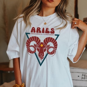 May include: White oversized t-shirt with a graphic of a red ram's head in a teal triangle. The text "Aries" is in a retro font above the ram and "Passionate + Confident" is below the ram.