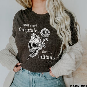 Peut inclure: T-shirt gris foncé chiné avec un motif de crâne et de rose blancs. Le texte sur le t-shirt est "I still read fairytales but for the villains now".