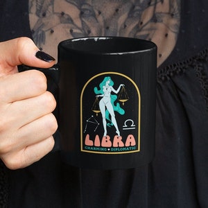 May include: Black ceramic mug with a white graphic of a woman with green hair standing on a scale. The graphic also includes the zodiac symbol for Libra and the text "Libra Charming + Diplomatic".