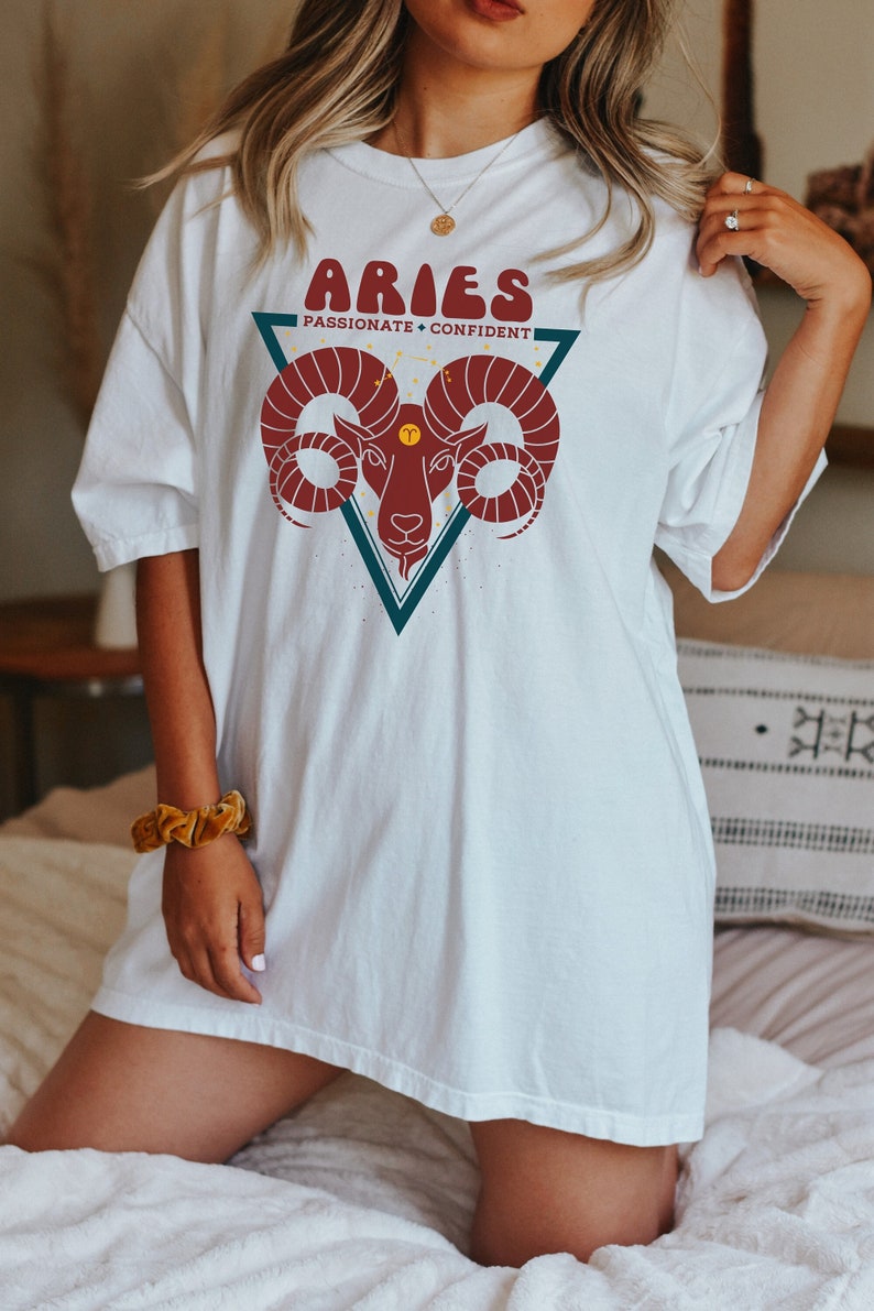 May include: White oversized t-shirt with a graphic of a red ram's head in a teal triangle. The text "Aries" is in a retro font above the ram and "Passionate + Confident" is below the ram.
