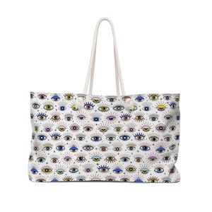 May include: A white canvas tote bag with a pattern of colorful eyes with eyelashes and rays of light. The bag has white rope handles.