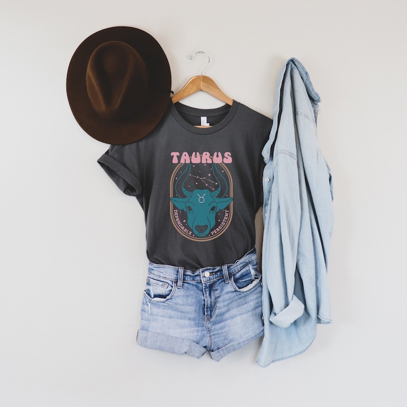K&ouml;nnte beinhalten: Ein graues T-Shirt mit einem Grafikdruck eines Bullenkopfes und dem Wort "Taurus" in rosa Buchstaben. Die Grafik enth&auml;lt auch die W&ouml;rter "dependable" und "persistent" in einem kreisf&ouml;rmigen Design.