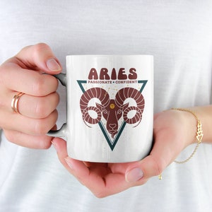 May include: White ceramic mug with a red and brown ram illustration inside a teal triangle. The text "Aries Passionate + Confident" is printed on the mug.