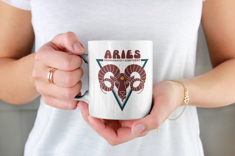 May include: White ceramic mug with a red and brown ram illustration inside a teal triangle. The text "Aries Passionate + Confident" is printed on the mug.