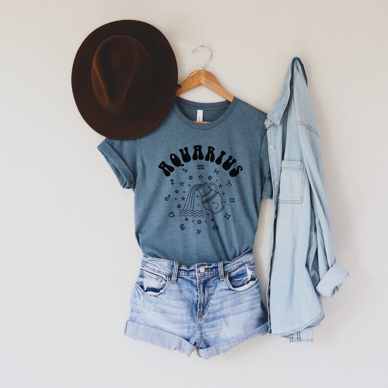 Puede incluir: Una camiseta azul con un gr&aacute;fico negro del signo astrol&oacute;gico de Acuario. El gr&aacute;fico presenta una jarra de agua estilizada que vierte agua y estrellas. La camiseta est&aacute; colgada en una percha de madera con un sombrero marr&oacute;n y una camisa vaquera azul.