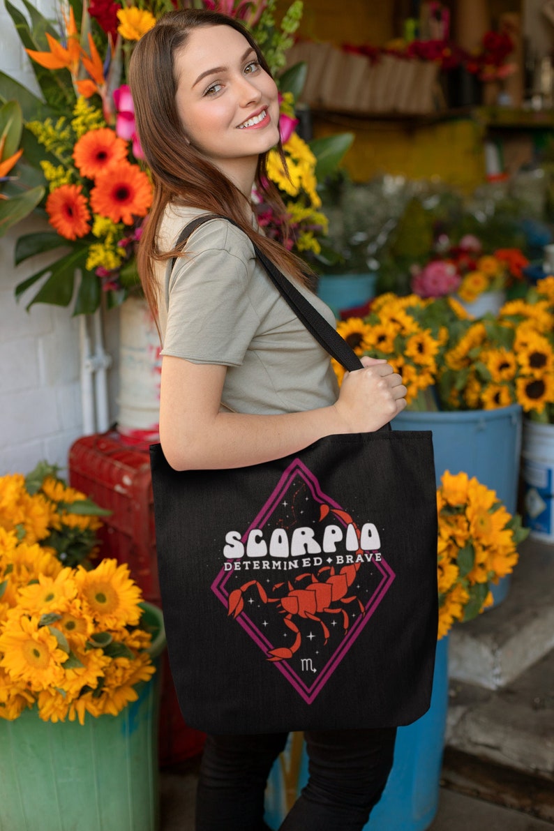 May include: Black tote bag with a Scorpio zodiac design. The design includes a red scorpion illustration and the words "Scorpio Determined + Brave". The bag has a long black strap and is held by a person in front of a flower shop.