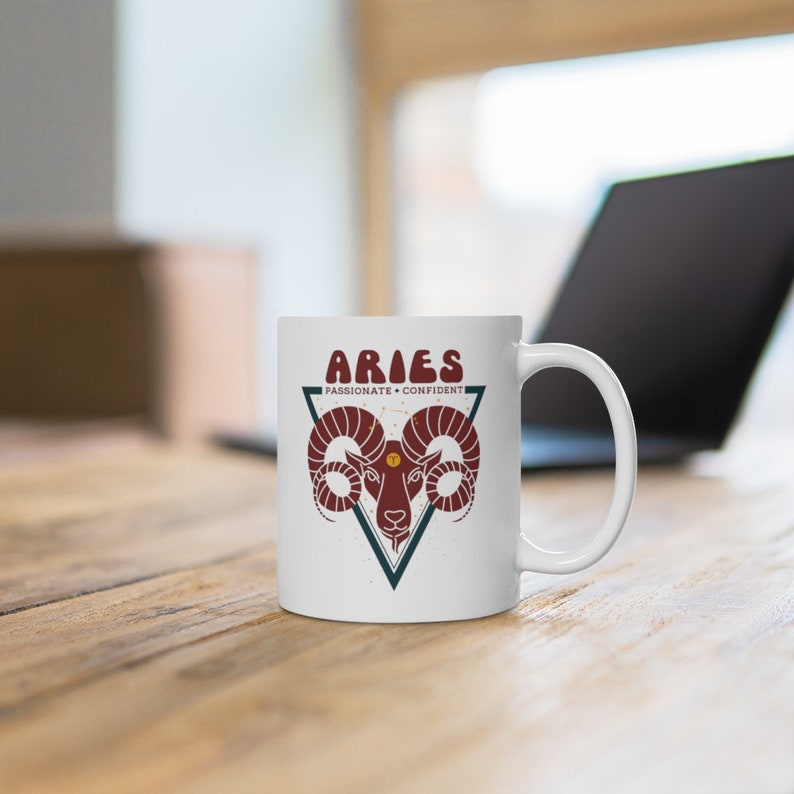 May include: White ceramic mug with a red and blue graphic of an Aries zodiac sign with the text "Aries Passionate + Confident".