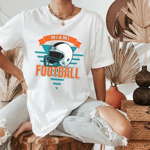 May include: White t-shirt with a teal and orange graphic design featuring a football helmet and the text "Miami Football".
