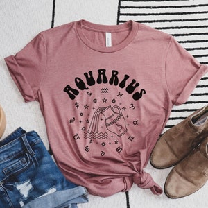 Puede incluir: Una camiseta rosa con un gr&aacute;fico negro del signo astrol&oacute;gico de Acuario. El gr&aacute;fico presenta una jarra de agua estilizada que vierte agua con estrellas y otros s&iacute;mbolos astrol&oacute;gicos que la rodean.