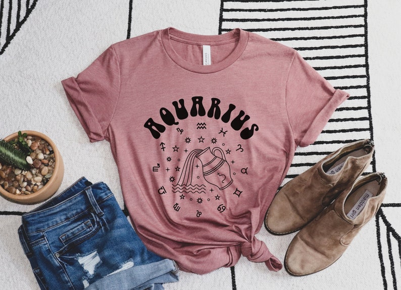 Puede incluir: Una camiseta rosa con un gr&aacute;fico negro del signo astrol&oacute;gico de Acuario. El gr&aacute;fico presenta una jarra de agua estilizada que vierte agua con estrellas y otros s&iacute;mbolos astrol&oacute;gicos que la rodean.