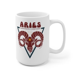 May include: White ceramic mug with a red and brown illustration of an Aries zodiac sign with the text "Aries Passionate + Confident" in a retro font.