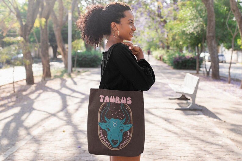 May include: A brown tote bag featuring the Taurus zodiac sign. The design includes the word "TAURUS" in pink, a blue bull illustration, and the words "DEPENDABLE" and "PERSISTENT". The bag is carried by a person.