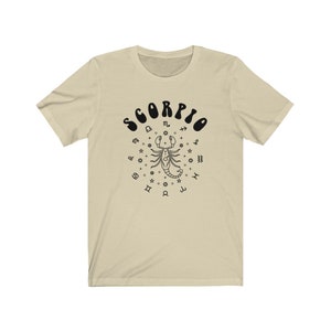 May include: A beige t-shirt with a black graphic of a scorpion surrounded by zodiac symbols and the word "SCORPIO" in a retro font.