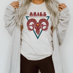 May include: A white t-shirt with a graphic of an Aries zodiac sign. The graphic is a red ram's head inside a blue triangle with the text "Aries" and "Passionate + Confident" in a retro font.