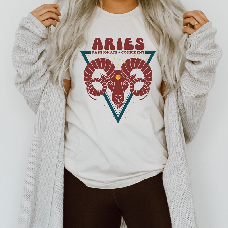 May include: A white t-shirt with a graphic of an Aries zodiac sign. The graphic is a red ram's head inside a blue triangle with the text "Aries" and "Passionate + Confident" in a retro font.