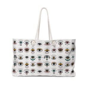 May include: A white canvas tote bag with a pattern of colorful eyes. The eyes are surrounded by a sunburst design and have various details like eyelashes, eyebrows, and teardrops. The bag has white rope handles.