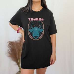 K&ouml;nnte beinhalten: Schwarzes T-Shirt mit einem Grafikdruck eines Bullenkopfes und dem Text "Taurus" in Pink. Die Grafik enth&auml;lt auch die W&ouml;rter "Dependable" und "Persistent" in Braun.
