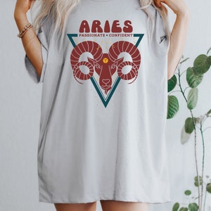 May include: A gray t-shirt with a graphic of an Aries zodiac sign. The graphic is a red ram's head inside a teal triangle with the words "Aries, Passionate + Confident" above it.