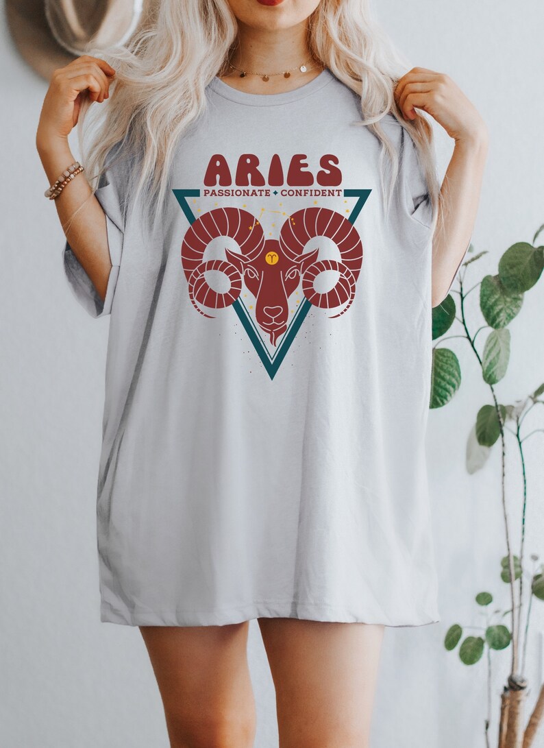 May include: A gray t-shirt with a graphic of an Aries zodiac sign. The graphic is a red ram's head inside a teal triangle with the words "Aries, Passionate + Confident" above it.