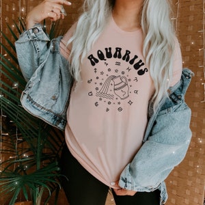 Puede incluir: Una camiseta rosa con un gr&aacute;fico negro del signo astrol&oacute;gico de Acuario. El gr&aacute;fico presenta una jarra de agua estilizada que vierte agua, rodeada de estrellas y s&iacute;mbolos del zod&iacute;aco.