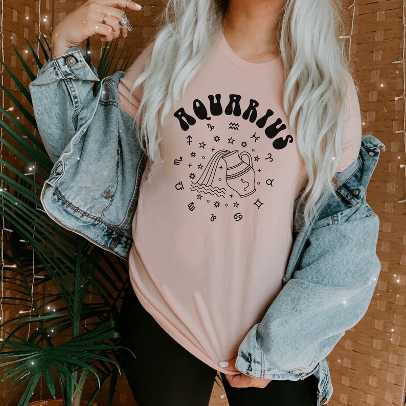 Puede incluir: Una camiseta rosa con un gr&aacute;fico negro del signo astrol&oacute;gico de Acuario. El gr&aacute;fico presenta una jarra de agua estilizada que vierte agua, rodeada de estrellas y s&iacute;mbolos del zod&iacute;aco.