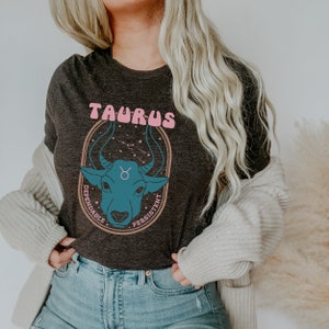 K&ouml;nnte beinhalten: Ein dunkelgraues T-Shirt mit einem Grafikdruck eines blauen Bullenkopfes mit rosa Schriftzug "Taurus" &uuml;ber dem Bullen und "Dependable" und "Persistent" auf beiden Seiten des Bullenkopfes.