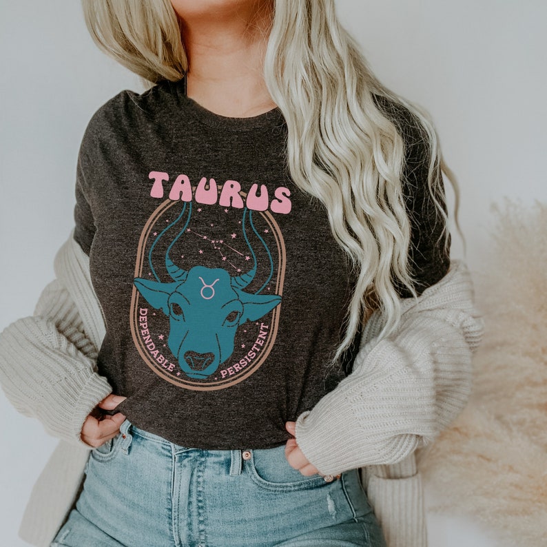 K&ouml;nnte beinhalten: Ein dunkelgraues T-Shirt mit einem Grafikdruck eines blauen Bullenkopfes mit rosa Schriftzug "Taurus" &uuml;ber dem Bullen und "Dependable" und "Persistent" auf beiden Seiten des Bullenkopfes.