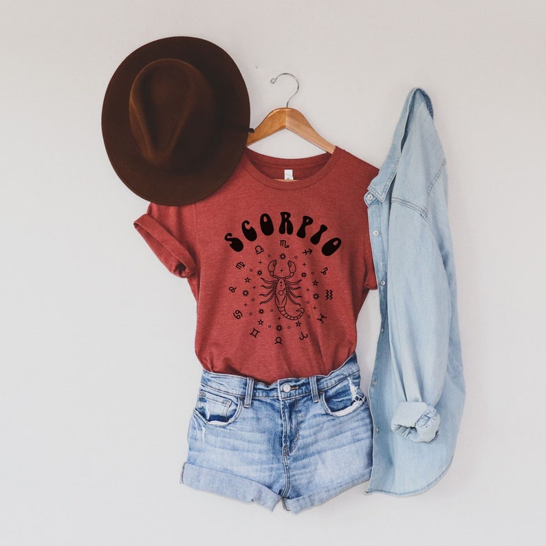 May include: A red t-shirt with a black graphic of the zodiac sign Scorpio. The graphic includes the word "Scorpio" in a retro font and a scorpion with astrological symbols around it.