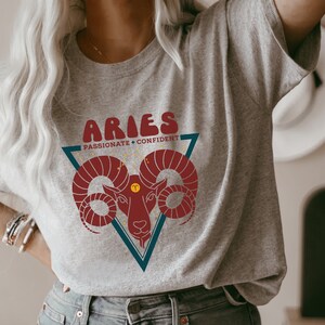 May include: A gray t-shirt with a red and blue graphic of an Aries zodiac sign. The graphic includes the text "Aries" in a retro font and the words "Passionate + Confident".