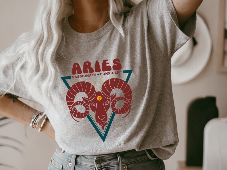 May include: A gray t-shirt with a red and blue graphic of an Aries zodiac sign. The graphic includes the text "Aries" in a retro font and the words "Passionate + Confident".