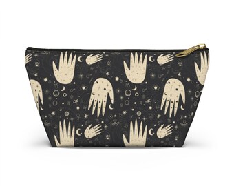 Könnte beinhalten: Schwarze Kosmetiktasche mit goldenem Reißverschluss. Die Tasche hat ein sich wiederholendes Muster aus beigefarbenen Händen, Sternen, Monden und Himmelsymbolen. Die Tasche ist für Make-up und andere Schönheitsprodukte konzipiert.