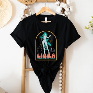 Może przedstawiać: Czarny T-shirt z grafiką przedstawiającą kobietę stojącą na wadze z nadrukiem "Libra Charming + Diplomatic" na przodzie.