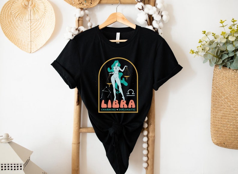 Może przedstawiać: Czarny T-shirt z grafiką przedstawiającą kobietę stojącą na wadze z nadrukiem "Libra Charming + Diplomatic" na przodzie.