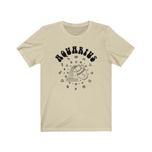 Puede incluir: Una camiseta beige con un gr&aacute;fico negro del signo astrol&oacute;gico de Acuario. El gr&aacute;fico presenta una jarra de agua estilizada que vierte agua y est&aacute; rodeada de s&iacute;mbolos del zod&iacute;aco.