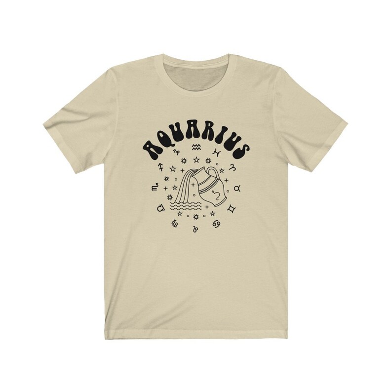 Puede incluir: Una camiseta beige con un gr&aacute;fico negro del signo astrol&oacute;gico de Acuario. El gr&aacute;fico presenta una jarra de agua estilizada que vierte agua y est&aacute; rodeada de s&iacute;mbolos del zod&iacute;aco.
