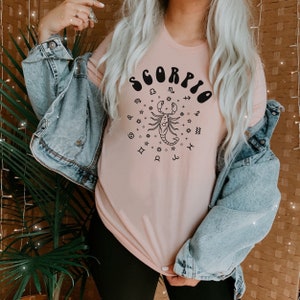 May include: A pink t-shirt with a black graphic of a scorpion and the word "Scorpio" in a retro font. The graphic also includes astrological symbols.