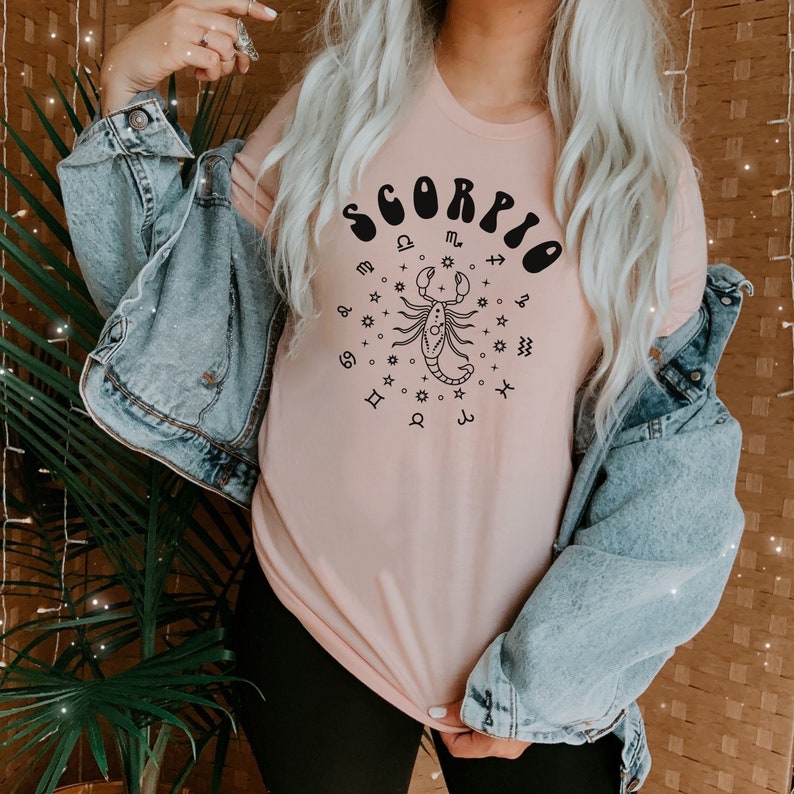 May include: A pink t-shirt with a black graphic of a scorpion and the word "Scorpio" in a retro font. The graphic also includes astrological symbols.