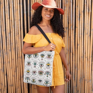 May include: A woman wearing a yellow dress and a wide-brimmed hat is holding a white tote bag with a colorful eye pattern. The bag has a black strap and is decorated with various eyes, stars, and other symbols.
