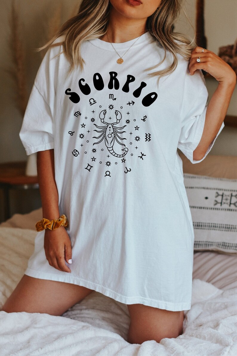 May include: White oversized t-shirt with a black graphic of the zodiac sign Scorpio. The graphic includes the word "Scorpio" in a retro font and a scorpion with astrological symbols.