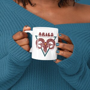 May include: White ceramic mug with a red and white illustration of an Aries zodiac sign with the text "Aries Passionate + Confident".