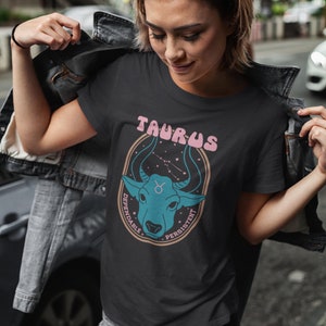 K&ouml;nnte beinhalten: Schwarzes T-Shirt mit einer blauen und pinkfarbenen Grafik eines Bullenkopfes mit Sternen und dem Text "Taurus" in pinkfarbenen Buchstaben. Die Texte "Dependable" und "Persistent" sind in Pink um den Bullenkopf herum geschrieben.