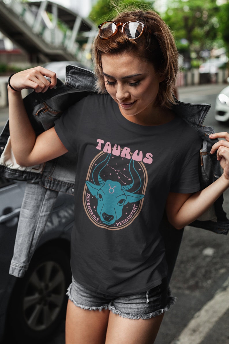 K&ouml;nnte beinhalten: Schwarzes T-Shirt mit einer blauen und pinkfarbenen Grafik eines Bullenkopfes mit Sternen und dem Text "Taurus" in pinkfarbenen Buchstaben. Die Texte "Dependable" und "Persistent" sind in Pink um den Bullenkopf herum geschrieben.