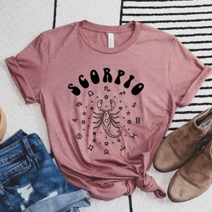 May include: A mauve t-shirt with a black graphic of a scorpion and the word "SCORPIO" in a retro font. The graphic is surrounded by zodiac symbols.