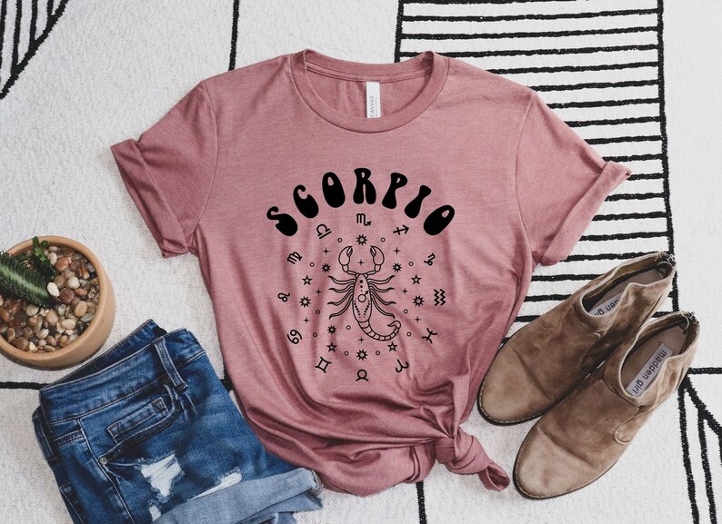 May include: A mauve t-shirt with a black graphic of a scorpion and the word "SCORPIO" in a retro font. The graphic is surrounded by zodiac symbols.
